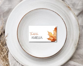 Puede incluir: Una mesa blanca con una pequeña tarjeta rectangular. La tarjeta presenta el texto "Thankful for AMELIA" en una fuente manuscrita, con una ilustración de hoja de arce en acuarela en tonos naranja y marrón.