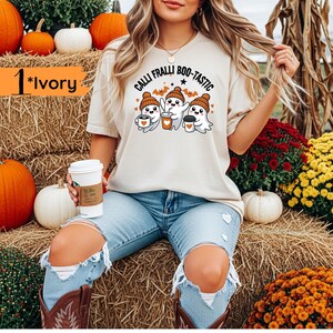May include: Ivory-colored t-shirt with the text "Calli Fralli Boo-tastic" and a graphic of three cartoon ghosts wearing hats and holding coffee cups. The shirt is worn by a person sitting on a hay bale surrounded by pumpkins and flowers.