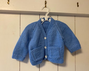 Puede incluir: Cárdigan de bebé de punto azul claro con dos bolsillos delanteros y botones blancos. El cárdigan está colgado de una percha blanca sobre un fondo de madera blanca. La prenda está hecha de un hilo suave y texturizado.