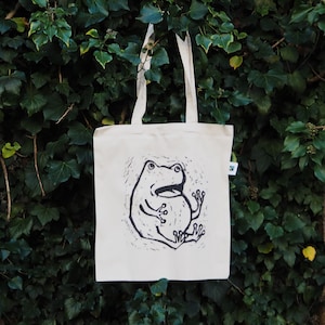 Peut inclure: Un sac fourre-tout en toile blanche avec une illustration en noir et blanc d'une grenouille sur le devant. La grenouille est assise les jambes croisées et a la bouche grande ouverte.