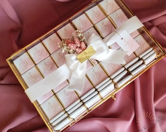 Può includere: Una scatola di vetro trasparente piena di piccoli quadrati di cioccolato rosa e bianco, ognuno con le lettere "SG" in lettere dorate. La scatola è legata con un nastro di raso bianco e decorata con un piccolo bouquet di fiori secchi.
