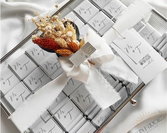 Può includere: Una scatola di vetro trasparente piena di piccole scatole bianche avvolte in un nastro bianco. Ogni scatola è etichettata con le iniziali "M & F". Un piccolo bouquet di fiori secchi è posto sopra le scatole. La scatola è etichettata "Sun Favors Wedding Gifts".