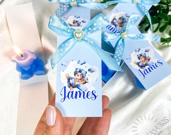 May include: Light blue favor boxes with a teddy bear pilot design, tied with blue polka dot ribbons and pearl accents. Each box has the name "James" printed on it. A blue candle is visible in the background.