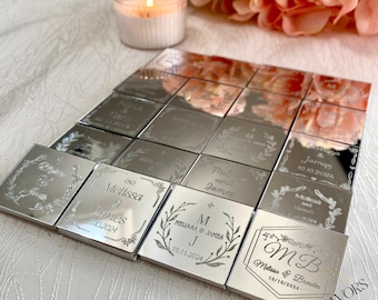 Op de afbeelding: Zilveren spiegelgeschenken met verschillende gegraveerde ontwerpen en datums. De geschenken hebben een ontwerp met een bloemkrans en de tekst "Melissa & James" en "Melissa & Brenden".