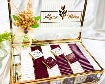 Può includere: Una scatola di vetro trasparente con una cornice dorata, piena di barrette di cioccolato. La scatola è decorata con una composizione floreale e i nomi "Alyssa" e "Mikey". Le confezioni di cioccolato sono bianche e bordeaux, con le iniziali "A M".
