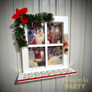 May include: A white framed Christmas decoration depicting a window scene with Santa Claus. The frame is decorated with a red poinsettia, green garland, and small baubles. A shelf with a festive pattern is below the window.