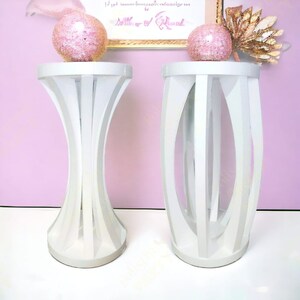 May include: Two white, decorative, open-work, cylindrical end tables with rounded tops. The table on the left has a flared base. The table on the right has a straight base.