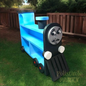 May include: Blue and black wooden toy train shaped like a bookshelf with shelves. The train has a face with a black moustache and a friendly smile. The text "Polistirolo Party" is visible on the bottom right of the image.