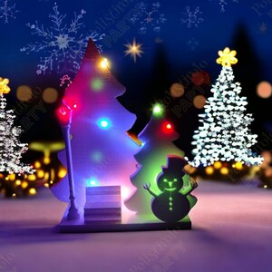 May include: A wooden Christmas scene with two illuminated Christmas trees, a snowman, and a street lamp. The trees are decorated with colourful lights, and the snowman wears a top hat. The background shows a snowy landscape with bokeh lights and snowflakes.