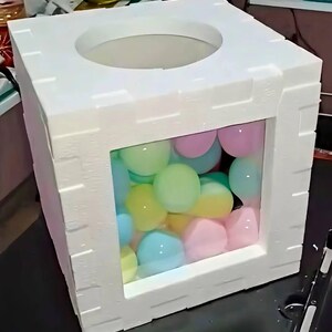 May include: A white, cube-shaped container with a circular opening on top and a rectangular window on the side. The window reveals colourful pastel-coloured balls inside. The word "PRIMA" is printed at the bottom.