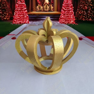 May include: A gold crown made of foam with a decorative fleur-de-lis on top. The crown is sitting on a white surface.