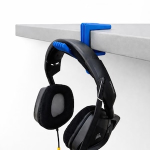 May include: A blue headphone holder is attached to a gray shelf, supporting a black headset. The headset has large earcups with black padding and a black headband. A yellow and black cable hangs down.