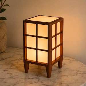 May include: A wooden cube-shaped table lamp with a grid design. The lamp has a warm, glowing light emanating from within. The lamp is supported by four short legs and sits on a marble-patterned surface. The lamp's dimensions are approximately 8 inches tall and 6 inches wide.