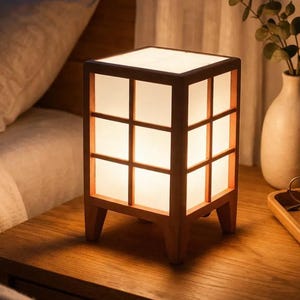 Puede incluir: Lámpara de noche cuadrada de madera con diseño de rejilla, que emite una luz suave y cálida. La lámpara está sobre una mesita de noche de madera, junto a un jarrón y una bandeja con gafas y un libro.
