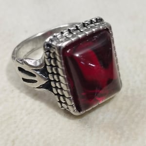 May include: A silver ring with a red gemstone set in a rectangular frame. The ring has a detailed design around the stone.