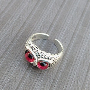 May include: A silver ring featuring an owl design. The owl has red eyes and intricate details.