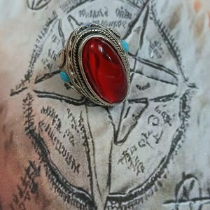 May include: A silver ring with a large, oval-shaped, red gemstone set in a decorative silver bezel. The ring has two small, turquoise gemstones on either side of the main stone.