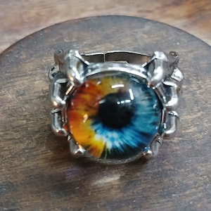 May include: A silver ring with a large, colorful eye design. The eye is blue, yellow, and orange with a black pupil.