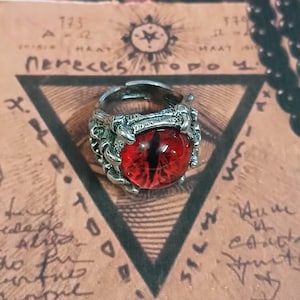 May include: A silver ring with a red eye and claw-like details. The ring is on a background with a triangle and writing.