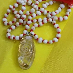 以下が含まれることがあります： 白と赤のビーズネックレスに、透明な楕円形のペンダントがぶら下がっています。ペンダントには金色のシンボルが描かれています。ペンダントは銀色のチェーンでつながっています。