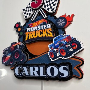 May include: A Hot Wheels Monster Trucks cake topper featuring a blue monster truck, a red number 3, chequered flags, and the name Carlos. The topper is set against a blue and orange background with a black glitter finish.