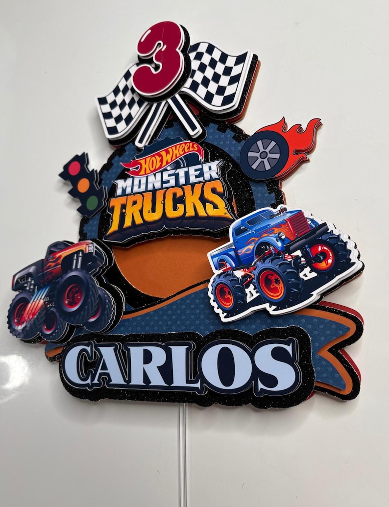 May include: A Hot Wheels Monster Trucks cake topper featuring a blue monster truck, a red number 3, chequered flags, and the name Carlos. The topper is set against a blue and orange background with a black glitter finish.