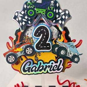 May include: A vibrant cake topper with monster trucks, chequered flags, and the number 2. The topper includes the name "Gabriel" in a decorative font, with flames and a skull and crossbones design. The theme is a birthday celebration.