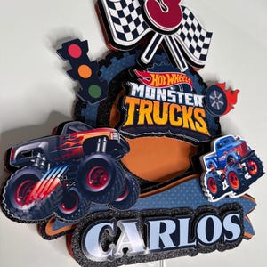 May include: A cake topper featuring a red, white and blue monster truck with the words "Hot Wheels Monster Trucks" and "Carlos". The topper also includes a chequered flag, a traffic light, and the number "3" in red.
