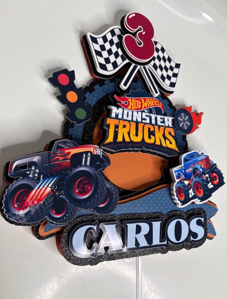 May include: A cake topper featuring a red, white and blue monster truck with the words "Hot Wheels Monster Trucks" and "Carlos". The topper also includes a chequered flag, a traffic light, and the number "3" in red.