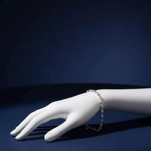 May include: A delicate silver bracelet with a repeating pattern of small, round gemstones. The bracelet is displayed on a white mannequin hand against a dark blue background. The jewelry is elegant and simple.
