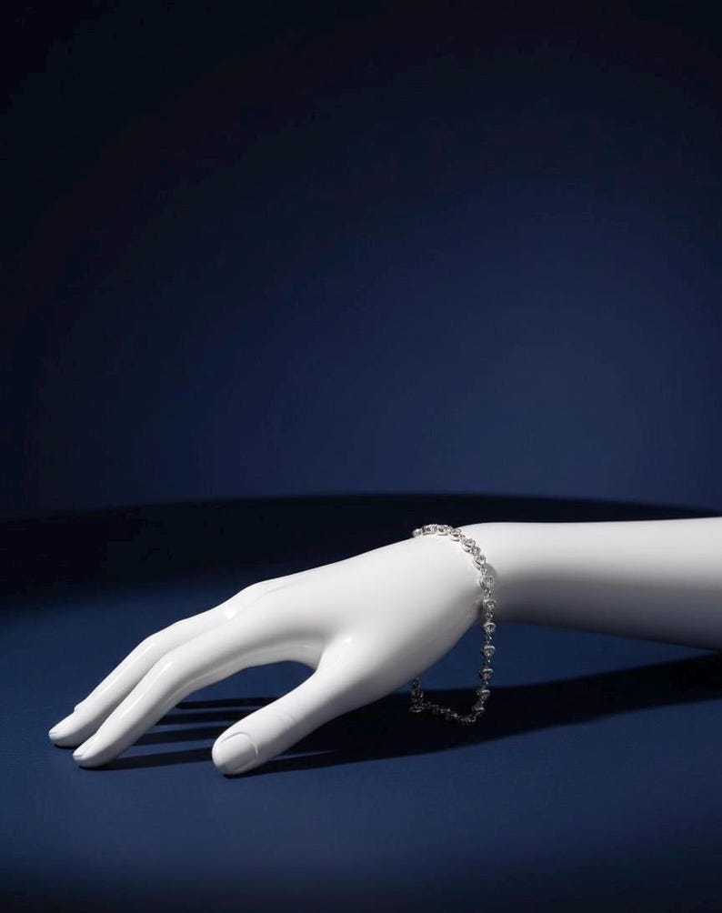 May include: A delicate silver bracelet with a repeating pattern of small, round gemstones. The bracelet is displayed on a white mannequin hand against a dark blue background. The jewelry is elegant and simple.