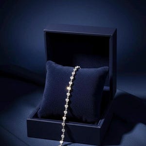 May include: A silver bracelet with heart-shaped gemstones rests on a dark blue velvet pillow inside a matching box. The box is open, revealing the bracelet's delicate design and sparkling stones. The background is a deep blue.
