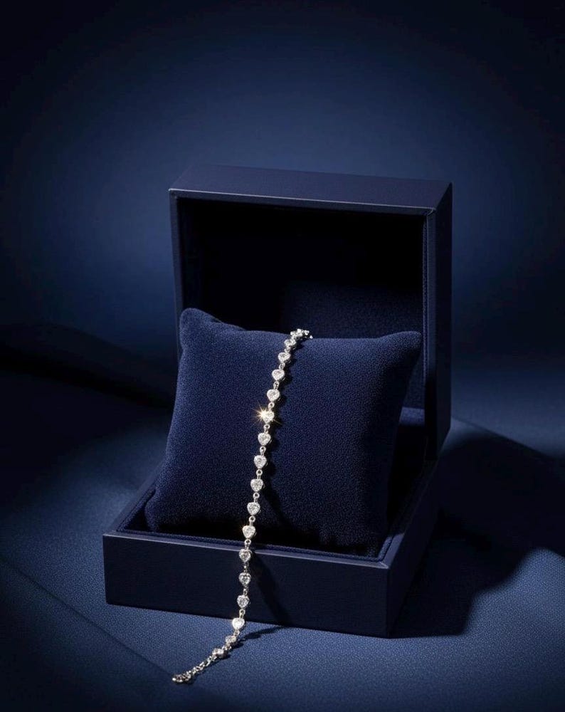 May include: A silver bracelet with heart-shaped gemstones rests on a dark blue velvet pillow inside a matching box. The box is open, revealing the bracelet's delicate design and sparkling stones. The background is a deep blue.