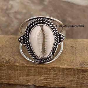 Può includere: Anello d'argento con una conchiglia di cauri bianca incastonata in una ghiera d'argento. L'anello ha una fascia attorcigliata.