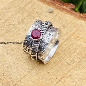 May include: A silver spinner ring featuring an oxidized finish with intricate floral engravings and a central, round, faceted deep red gemstone. The band includes a textured, ladder-like pattern section. The text LuckyJewellersGifts is visible.