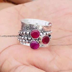 May include: A silver spinner ring featuring an oxidized floral pattern and three round, faceted, deep pink ruby gemstones. The band includes beaded silver wire detailing and the text LuckyJewellersGifts is visible on the side of the metal surface.