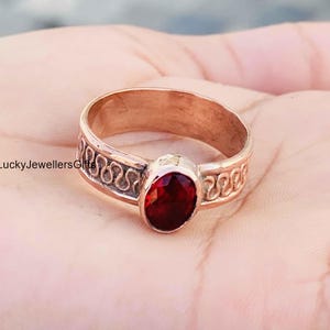 Könnte beinhalten: Ein Kupferring mit einem zentralen, oval geschliffenen, tiefroten Edelstein. Das Band zeigt ein durchgehendes, geprägtes Wellenmuster entlang seines Umfangs. Das Metall hat einen warmen Kupferton und der Stein ist facettiert und poliert.