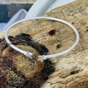 May include: A silver-toned open cuff bracelet featuring two small, five-pointed star charms at each end of the band. The metal has a smooth, polished finish, and the design is circular with a gap between the two star-shaped terminals.