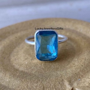 Puede incluir: Anillo de plata con una piedra preciosa azul rectangular facetada. La piedra está engastada en una banda de plata pulida. El texto "LuckyJewellersGifts" es visible en la imagen.