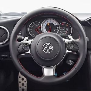 May include: Black leather steering wheel with red stitching, silver accents, and integrated controls. The center features a logo with the letters "DS". The dashboard displays gauges and warning lights.