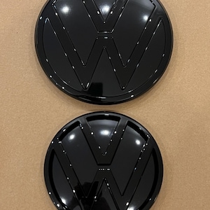 May include: Two black Volkswagen emblems. The larger emblem is approximately 7.5 inches in diameter and the smaller emblem is approximately 5.5 inches in diameter.