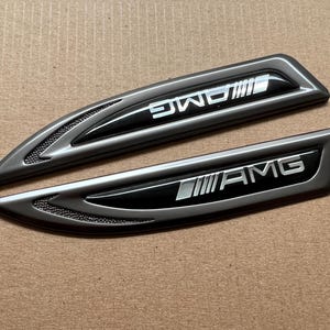 以下が含まれることがあります： 黒いアクセントが付いた銀色の車のエンブレムが2つ。上のエンブレムには「AMG」と書いてあり、下のエンブレムには「AMG」と書いてあります。