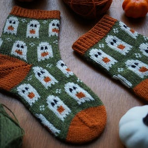 Puede incluir: Un par de calcetines de punto con tem&aacute;tica de Halloween. Los calcetines son de color verde oliva con un estampado de fantasmas blancos con barrigas de calabaza naranja. Los pu&ntilde;os y los dedos son de color naranja. Los calcetines se exhiben sobre una superficie de madera.