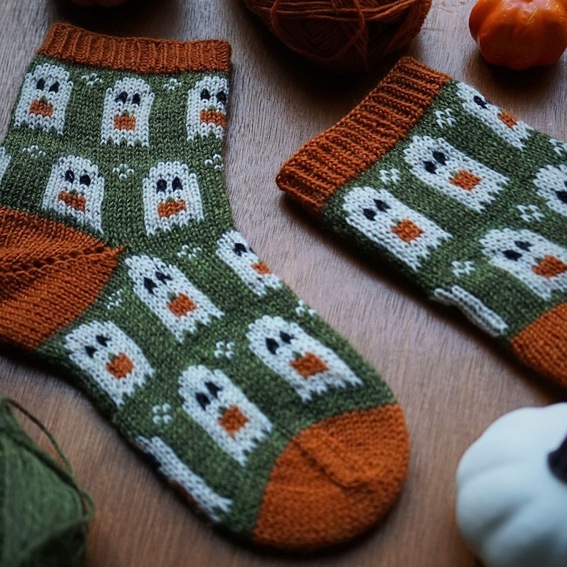 Puede incluir: Un par de calcetines de punto con tem&aacute;tica de Halloween. Los calcetines son de color verde oliva con un estampado de fantasmas blancos con barrigas de calabaza naranja. Los pu&ntilde;os y los dedos son de color naranja. Los calcetines se exhiben sobre una superficie de madera.