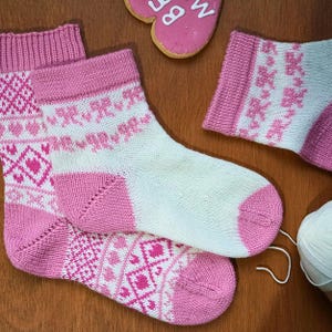 Könnte beinhalten: Handgestrickte Socken in Weiß und Rosa mit Herz- und Schleifenmustern. Die Socken haben rosa Fersen, Zehen und Bündchen. Ein Knäuel weiße Wolle und ein herzförmiger Keks sind ebenfalls zu sehen.