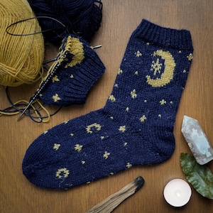 Puede incluir: Un par de calcetines azules de punto con un patrón de media luna amarilla y estrellas. Los calcetines están sobre una superficie de madera con ovillos de lana, una vela y un cristal.