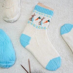Könnte beinhalten: Eine weiße Stricksocke mit blauer Ferse und Spitze sowie einem blauen Bündchen. Die Socke zeigt ein Muster aus braunen Hasen und orangenen Karotten. Stricknadeln und Knäuel aus weißer und blauer Wolle sind ebenfalls zu sehen.