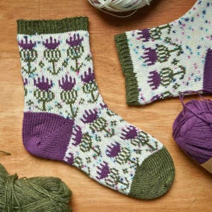 May include: A knitted sock featuring a repeating purple and green thistle pattern on a cream background, with a solid olive green ribbed cuff, a solid purple heel, and a solid olive green toe, accompanied by matching yarn skeins.