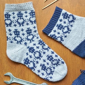 May include: A pair of knitted socks in light gray with a navy blue pattern. The socks feature a repeating floral design and have navy blue cuffs and heels. The socks are displayed on a wooden surface with knitting needles and a wrench.