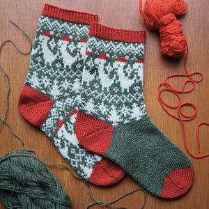 May include: A pair of knitted socks with a festive design. The socks feature a green and white pattern with reindeer and snowflakes, red heels, toes, and cuffs. The socks are displayed on a wooden surface with balls of yarn.
